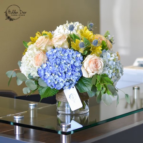 Blue hydrangeas and cream roses with yellow alstroemeria in glass vase by Raleigh floral design